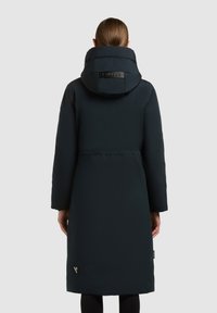 Femeie îmbrăcată cu un palton lung, negru, căptușit, cu guler înalt și glugă, văzută din spate pe un fundal simplu de culoare deschisă.