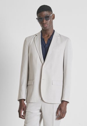 Man draagt een lichtbeige pak, marineblauw overhemd en donkere zonnebril, staat tegen een effen witte achtergrond.