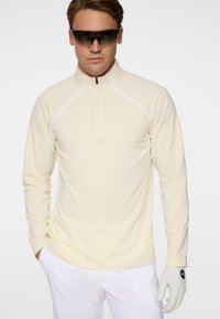 Homme portant un haut zippé manches longues beige, un pantalon blanc, un gant de golf blanc et des lunettes de soleil de sport foncées sur fond blanc.