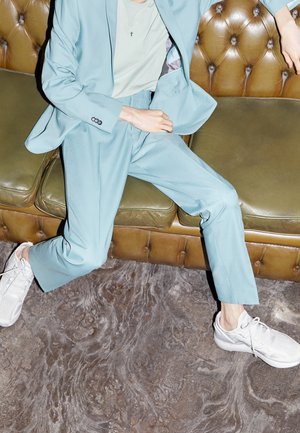 Costume turquoise à coupe ajustée, associé à un t-shirt vert clair et des baskets blanches, assis sur un canapé en cuir marron.