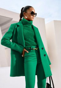 Groene wollen jas met zwarte knopen, gestructureerde coltrui en bijpassende broek. Accessoires omvatten een riem met een gouden accent en pareloorbellen.