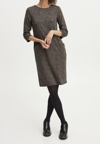 Ruskean leopardiprintin mekko, jossa on kolmen neljänneksen hihat, pyöreä kaula-aukko, taskut ja suora leikkaus, yhdistettynä mustiin leggingsseihin ja tukevasiin kenkiin.