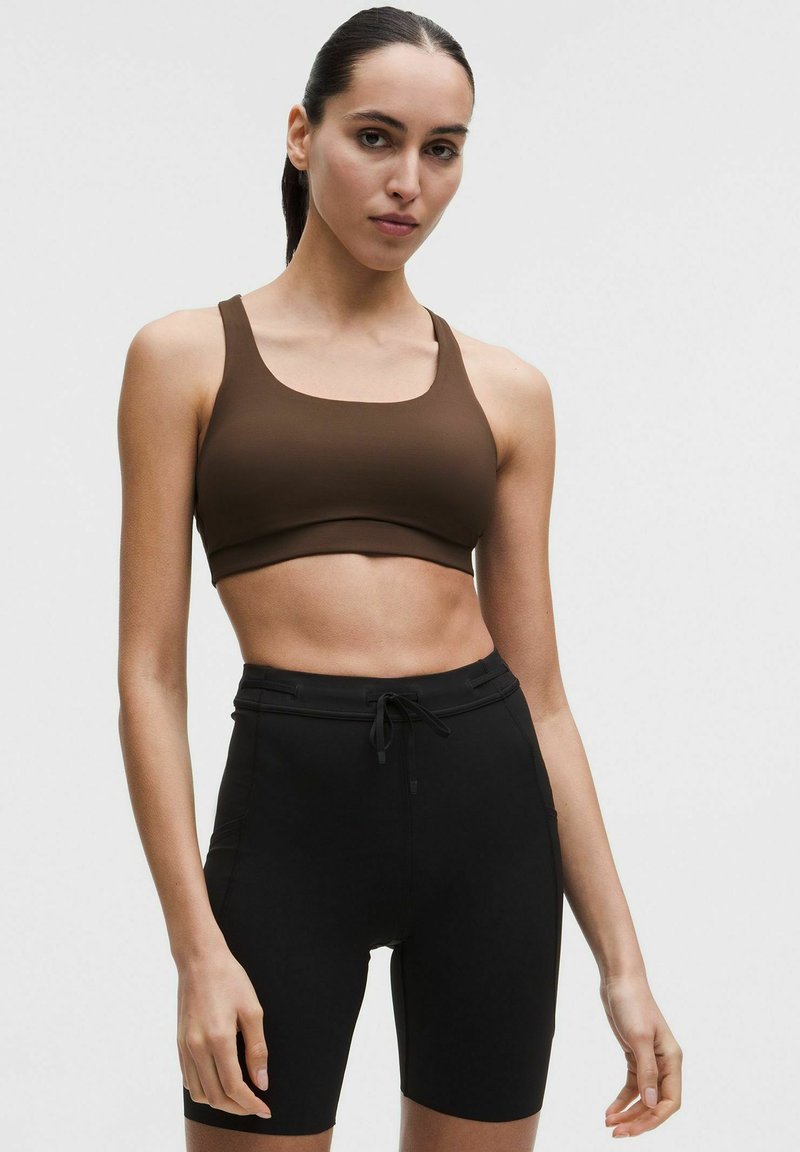 Frau mit dunklem Haar im Pferdeschwanz, die einen braunen Sport-BH und schwarze, hoch taillierte Radlerhosen vor einfachem Hintergrund trägt.