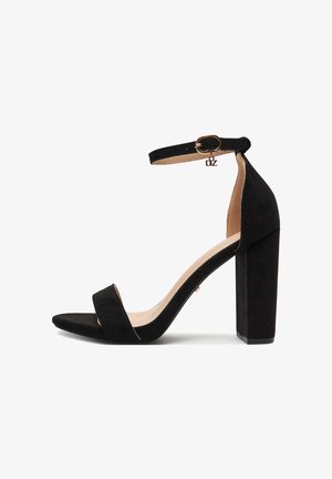 Czarna zamszowa sandałka na wysokim, szerokim obcasie, z odkrytymi palcami, paskiem wokół kostki i złotym klamrą. Gładka wewnętrzna wyściółka z minimalnymi zdobieniami.