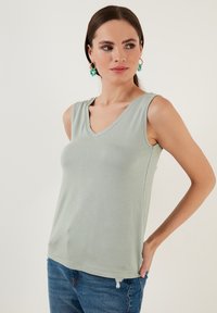 Svetlo zelena brezrokavna majica z V-izrezom iz mehkega materiala, ki ima oprijet kroj in čist rob. V kombinaciji z modrimi raztrganimi denim hlačami.