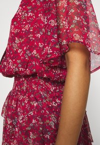 Robe rouge en tissu transparent à motifs floraux, taille froncée et manches volantes à côté d'un bras brun visible.