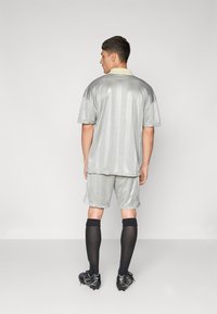 Camisola e calções desportivos prateados com padrões texturizados, meias de joelho pretas e botas pretas, vistos por trás.