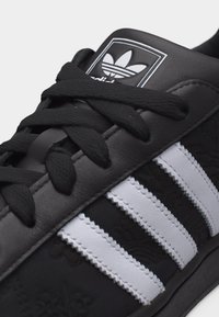 Μαύρο αθλητικό παπούτσι Adidas με λευκές τρεις ρίγες, μαύρα κορδόνια και ανάγλυφο λουλουδάτο σχέδιο στην πλαϊνή επιφάνεια.