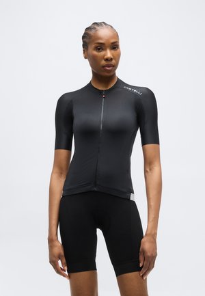 Γυναίκα που φοράει εφαρμοστό μαύρο ποδηλατικό τζέρσεϊ κοντών μανικιών και μαύρο ποδηλατικό σορτς, όρθια μπροστά σε ένα απλό ανοιχτό φόντο.