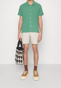 Grön kortärmad skjorta med knappar, beige shorts, bruna ankelsockor och beige sneakers. Håller en mönstrad tote-väska med svarta accenter.