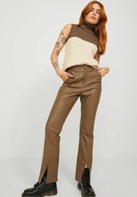 Jersey de cuello alto marrón y crema con un patrón de zigzag, combinado con pantalones de cuero sintético marrón ajustados con aberturas frontales y botas negras.