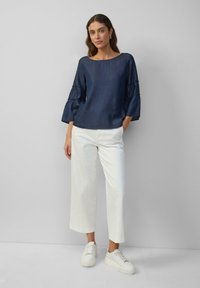 Marineblaue, strukturierte Bluse mit breiten, gerüschten Ärmeln, kombiniert mit weißen, taillierten gerade geschnittenen Hosen und weißen Sneakers.
