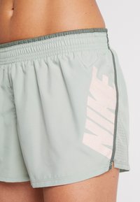 Primer plano de una persona que lleva pantalones cortos deportivos Nike de color verde claro con una cinturilla elástica y un gran logo de Nike en color rosa pálido en el lado.