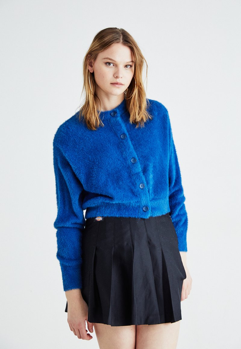 Cardigan blu in peluche con collo rotondo e sei bottoni blu, abbinato a una gonna nera a pieghe. Texture morbida, silhouette aderente e design casual.