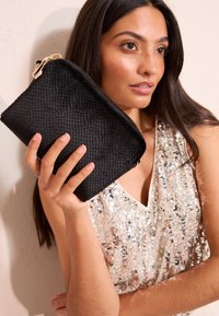 Clutch nero con texture e chiusura con zip, tenuta da una persona. Sullo sfondo si vede una maglia scintillante in tonalità beige e oro.