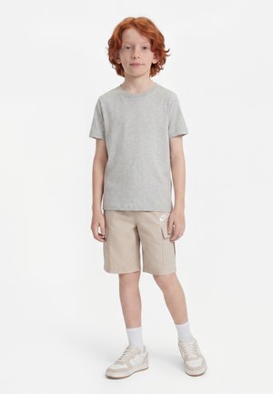 Băiat roșcat stând, purtând un tricou gri deschis, pantaloni scurți cargo Nike bej, șosete albe și adidași bej pe fundal alb.