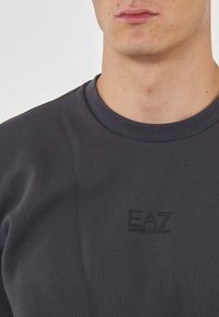 T-shirt grigio scuro a maniche corte realizzato in tessuto morbido, con scollatura rotonda e un sottile logo EA7 stampato sul lato sinistro del petto.