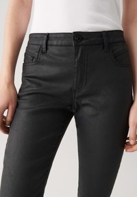 Donna in pantaloni di pelle neri attillati e una maglietta bianca a coste senza maniche, con le mani rilassate ai lati.