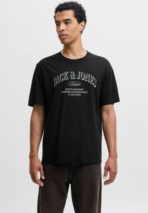 Mand iført sort Jack & Jones T-shirt med hvid tekst, mørke jeans og en sølvkæde, som kigger til siden mod en ensfarvet baggrund.