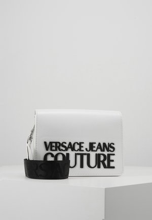 Bílá crossbody kabelka Versace Jeans Couture s výrazným černým logem a odnímatelným černým popruhem ležící na bílém povrchu.