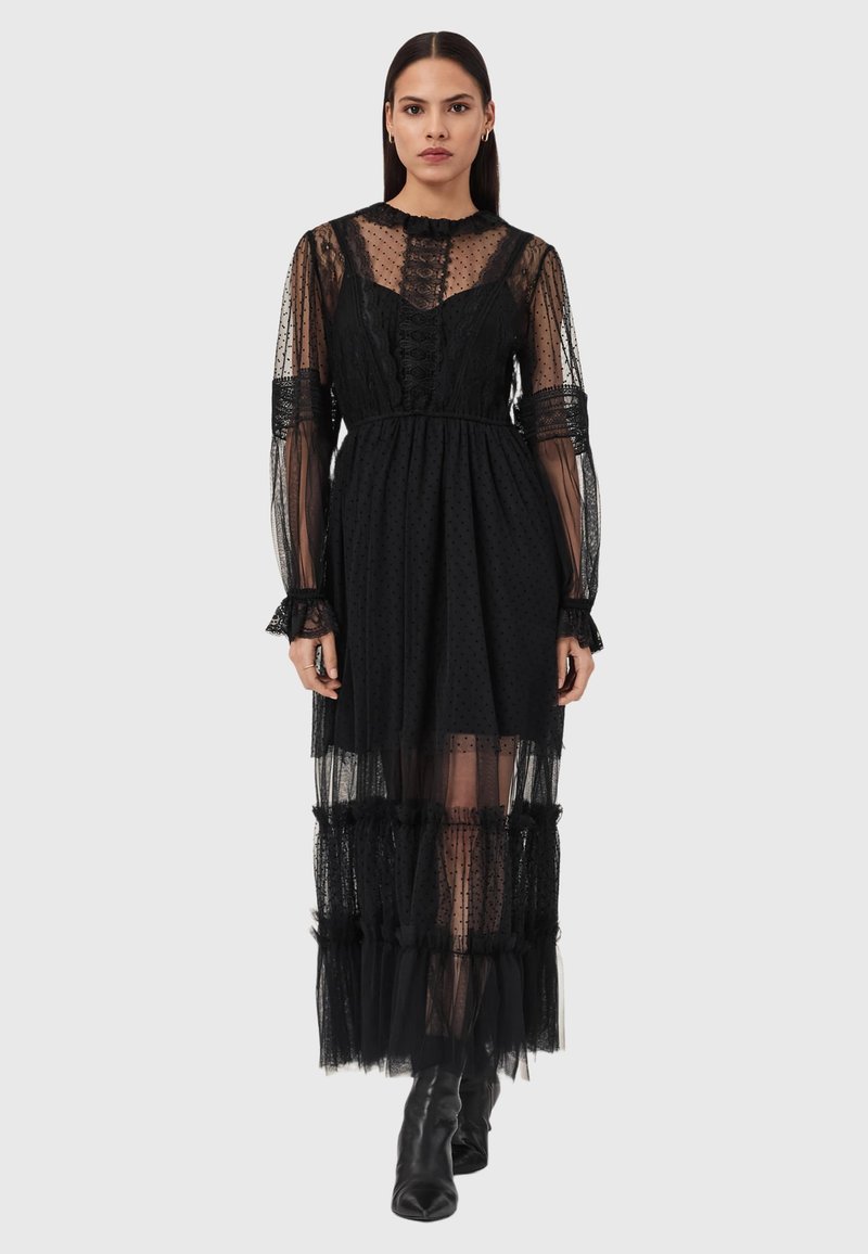 Μαύρο φόρεμα από δίχτυ με  υφή πουά, ψηλό λαιμό, μακριά μανίκια με βολάν και πολυεπίπεδα στρώματα τούλι. Διαθέτει εφαρμοστή κορυφή και φαρδιά φούστα.