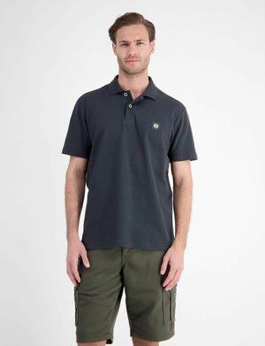 Mand iført en sort polo med krave og grønne cargoshorts mod en ensfarvet hvid baggrund.