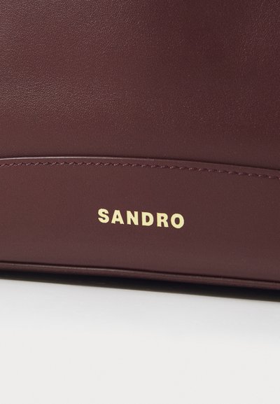 Burgundy vetokäsilaukku, jossa on sileä tekstuuri ja kultainen "SANDRO"-logo painettuna sivulle. Siistit linjat ja minimalistinen muotoilu.