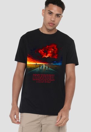 Young man wearing black t-shirt with Stranger Things logo and graphic of four kids riding bikes on a road under a fiery red and dark sky.