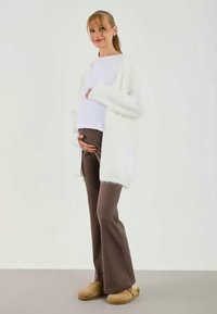 Cardigã branco tricotado sobre uma blusa branca, combinado com calças castanhas com textura em canalé e calçado bege. A modelo segura uma barriga de grávida, sorrindo.