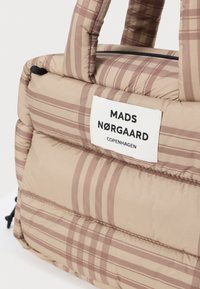 Beżowa torba z wypełnieniem, w brązowy wzór w kratę. Posiada podwójne uchwyty i białą metkę z czarnym tekstem. Zapinana na zamek błyskawiczny u góry.