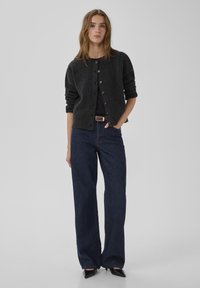 Cardigan de punto gris oscuro con botones, combinado con jeans de mezclilla oscuros de pierna ancha. Tacones negros de punta afilada completan el conjunto.