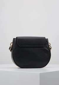 Borsa a tracolla in pelle nera con texture, chiusura a pattina e hardware in tonalità oro, esposta su un piedistallo bianco contro uno sfondo neutro.