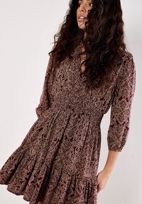 Frau mit langen, lockigen, dunklen Haaren, die ein weinrotes Kleid mit beigefarbenen, filigranen Blumenmustern und Dreiviertelärmeln trägt und seitlich posiert.