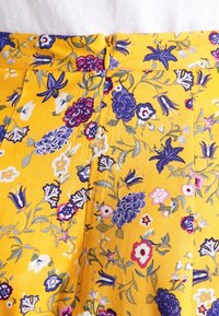 Tessuto floreale giallo con fiori viola, rosa e bianchi. Presenta una chiusura con zip in vita e una texture liscia e leggera.