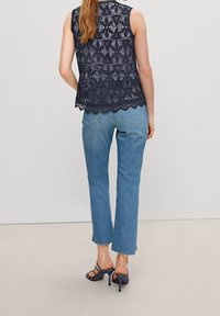 Marineblaues Spitzenoberteil mit strukturierten Mustern und gewelltem Saum; kombiniert mit hellblauen geraden Jeans und dunklen Riemchenabsätzen.