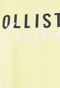 Żółta bawełniana koszulka z czarnymi literami w bloku, na której widnieje napis "HOLLISTER", a poniżej w kursywie znajduje się biały tekst "California".