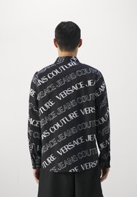 Svart skjorta med ett upprepande vitt mönster av "Versace Jeans Couture", långa ärmar och en något avslappnad passform. Strukturerad tygdetalj synlig.