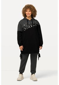 Schwarzer und grauer übergroßer Hoodie mit dem Text "SMILE", kombiniert mit grauen Jogginghosen. Kapuzen-Design, weicher Stoff und seitliche Bindetails. Weiche Sneakers.