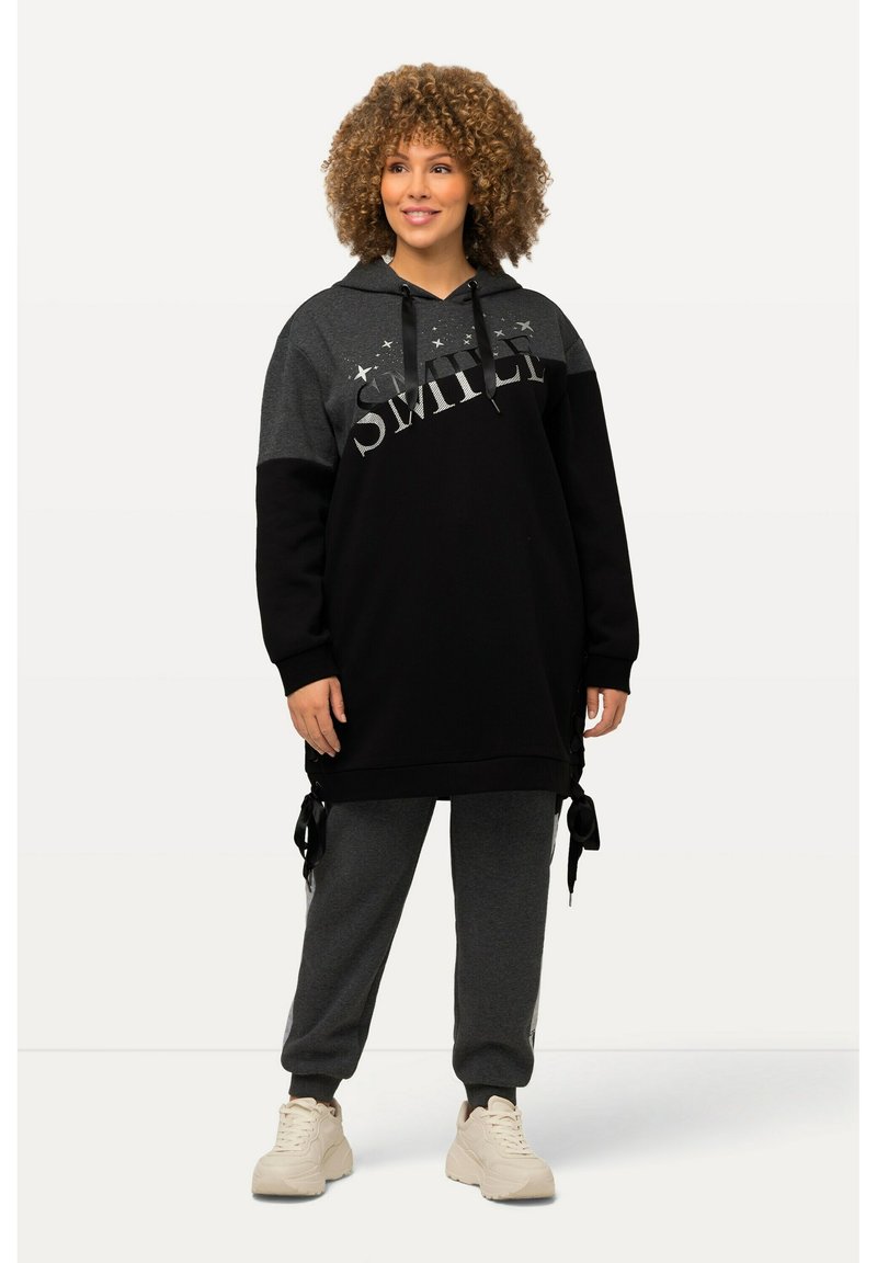 Schwarzer und grauer übergroßer Hoodie mit dem Text "SMILE", kombiniert mit grauen Jogginghosen. Kapuzen-Design, weicher Stoff und seitliche Bindetails. Weiche Sneakers.