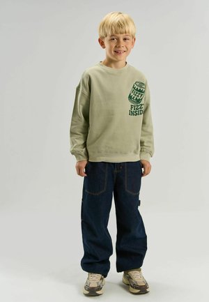 Jeune garçon blond souriant portant un sweat-shirt beige avec un motif canette de soda, jean foncé et baskets beiges, debout sur un fond uni.