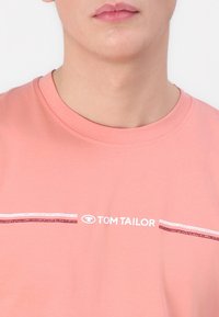 Šviesiai rožinė medvilninė marškinėlių su apvaliu iškirpimu, trumpomis rankovėmis ir grafiką, kurioje užrašyta "TOM TAILOR" ir dvi horizontalios juostos baltos ir kaštonų spalvų.
