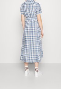 Žena v modro-bílé kárované midi šatech, bílých teniskách a károvaných kotníkových ponožkách, stojící proti jednoduchému pozadí, zády k divákovi.