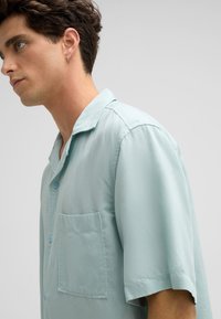 Kurzärmliges, hellblaues Hemd aus weichem Stoff, mit Kragen, Brusttasche und Knopfverschluss. Glatte Textur mit lockerer Passform.