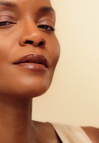 Primo piano del volto di una donna con pelle liscia, sottotoni beige morbidi, labbra marroni opache e trucco per occhi minimale su uno sfondo chiaro.