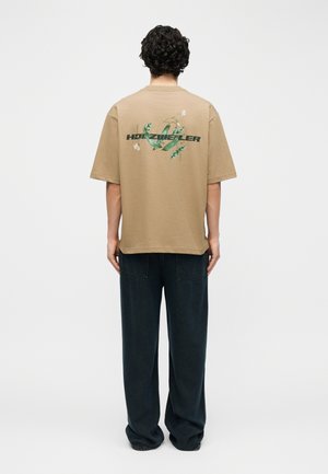 Άτομο με σγουρά μαλλιά στέκεται στραμμένο προς τα πίσω, φοράει μπεζ μπλουζάκι με πράσινο σχέδιο κληματαριάς και κείμενο, συνδυασμένο με φαρδιά σκούρα παντελόνια.