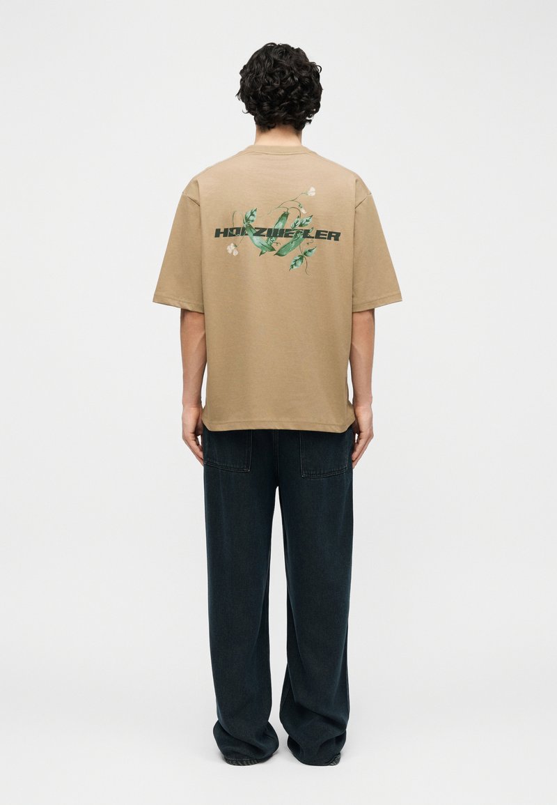 Άτομο με σγουρά μαλλιά στέκεται στραμμένο προς τα πίσω, φοράει μπεζ μπλουζάκι με πράσινο σχέδιο κληματαριάς και κείμενο, συνδυασμένο με φαρδιά σκούρα παντελόνια.