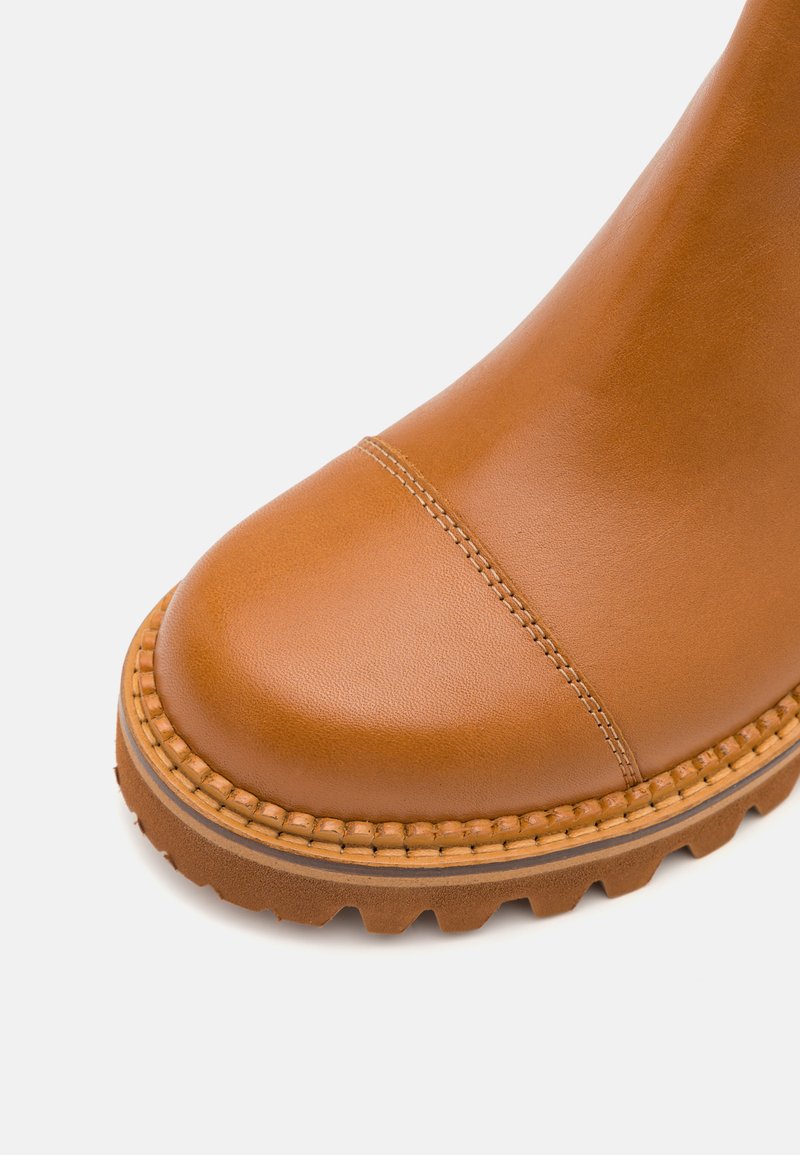 Stivale alla caviglia in pelle marrone chiaro con punta arrotondata, cuciture visibili, suola in gomma testurizzata e dettagli intrecciati attorno alla base.