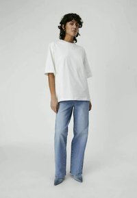 T-shirt bianco oversized con maniche corte, abbinato a jeans azzurri chiari. Modella che indossa tacchi a punta azzurri chiari, in piedi su uno sfondo neutro.