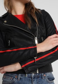 Giacca da moto in pelle nera con strisce rosse sulle maniche, chiusura frontale con zip e tasche laterali, caratterizzata da una texture trapuntata sulle spalle.