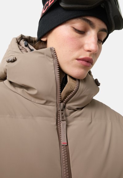 Beżowa kurtka puchowa z wysokim kołnierzem i zamkiem z przodu. Charakteryzuje się gładką fakturą, elementami regulowanymi oraz metką na zamku.